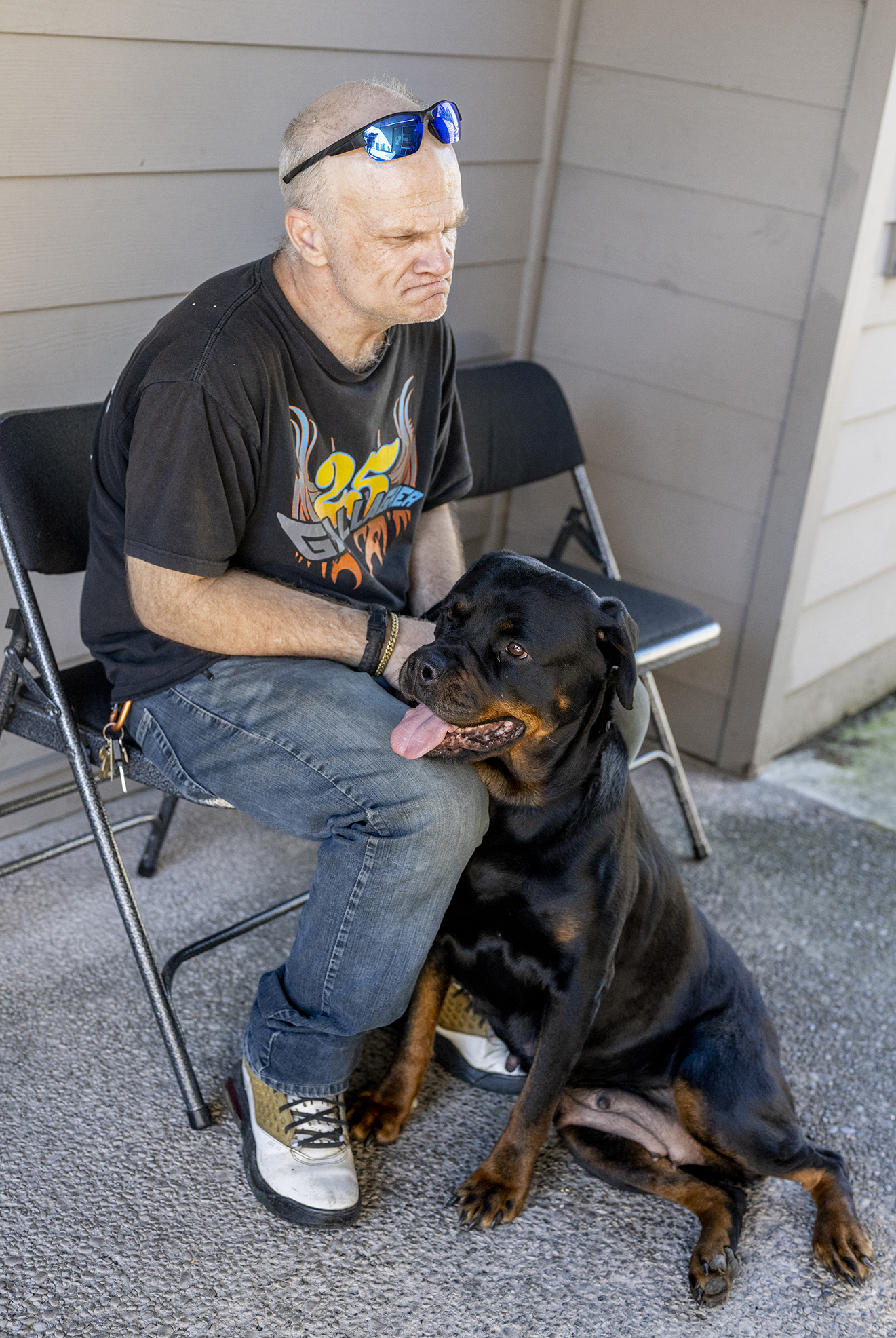 Steve sits in a plastic folding chair with his dog, Mamas, a black Rottweiler resting her head on his knee and leaning against him.