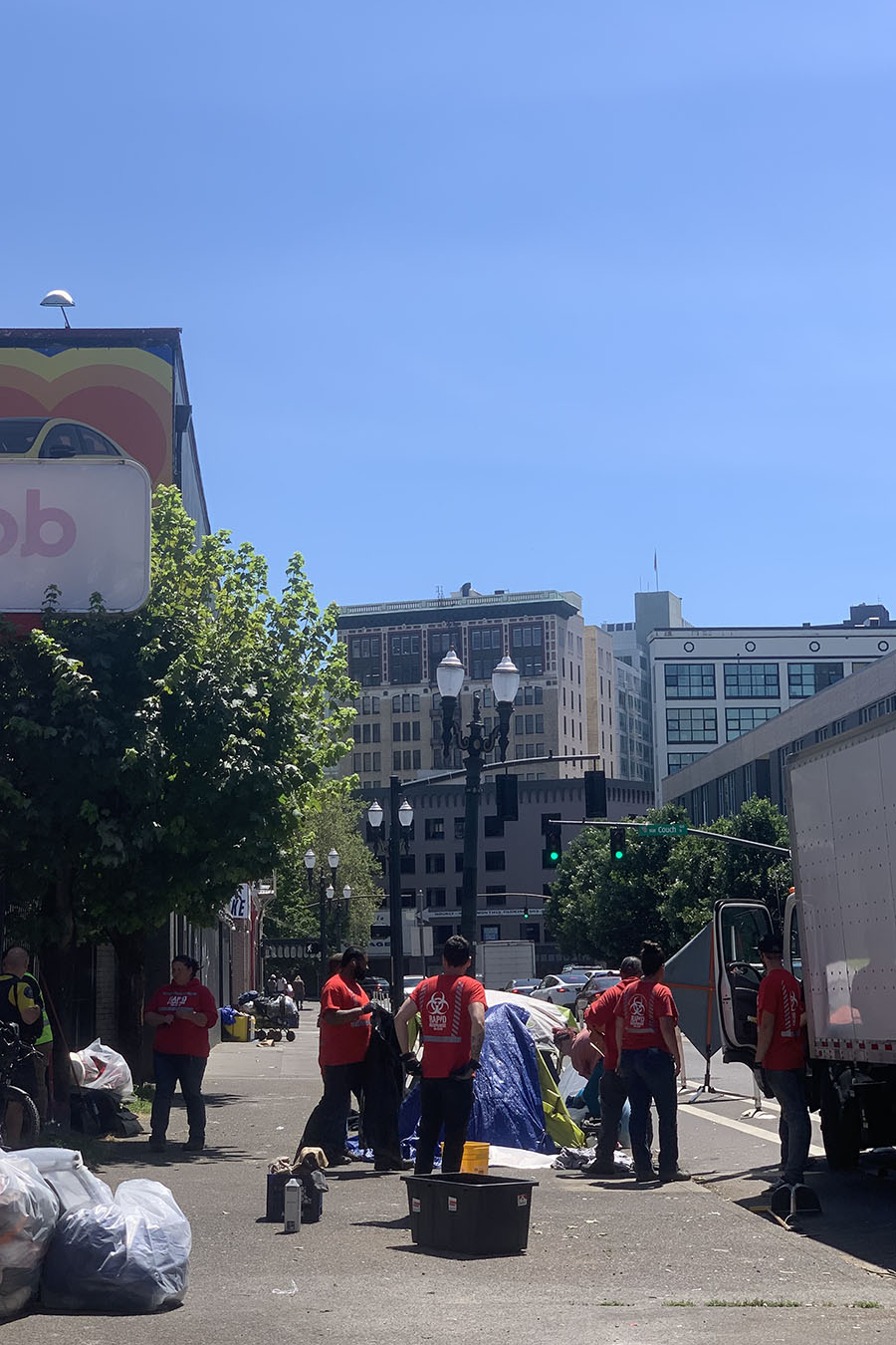 A group of workers in red stands on the sidewalk. On the left side of the sidewalk are plastic bags filled with posessions. In the background are blue skies and buildings.